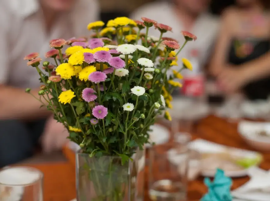 Ramo de flores coloridas, incluyendo amarillas, moradas y blancas, en un jarrón sobre una mesa con vasos y platos. Fondo desenfocado con personas en una reunión social.
