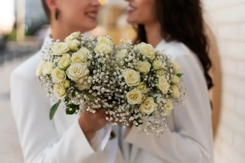 Pareja sonriente sosteniendo un ramo de rosas blancas y flores de gypsophila, vestidas con trajes blancos, en un entorno urbano. Ideal para bodas y celebraciones románticas.