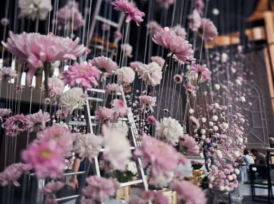 Decoración floral suspendida en un evento, con flores en tonos rosa y blanco colgando de hilos. Se observan escaleras y personas trabajando en el fondo.