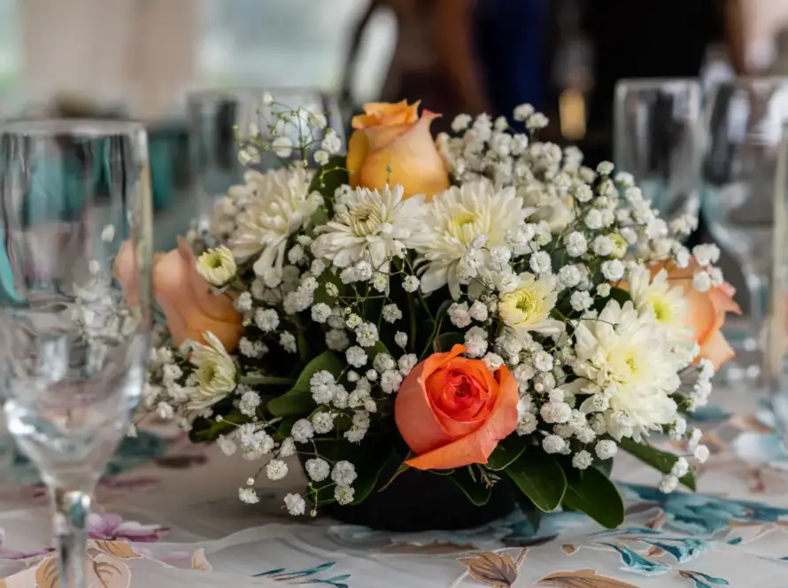 Centro de mesa floral con rosas naranjas y blancas, margaritas y flores de gypsophila, decorando una mesa elegante con copas de cristal. Ideal para eventos y celebraciones.