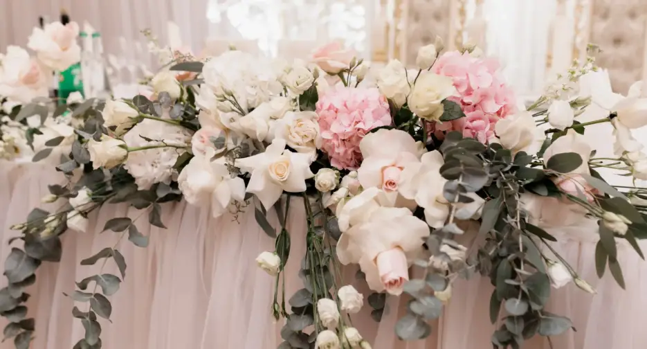 Arreglos florales elegantes con rosas blancas, rosas rosadas, hortensias y hojas de eucalipto sobre una mesa decorada con tela blanca, ideales para eventos y bodas.